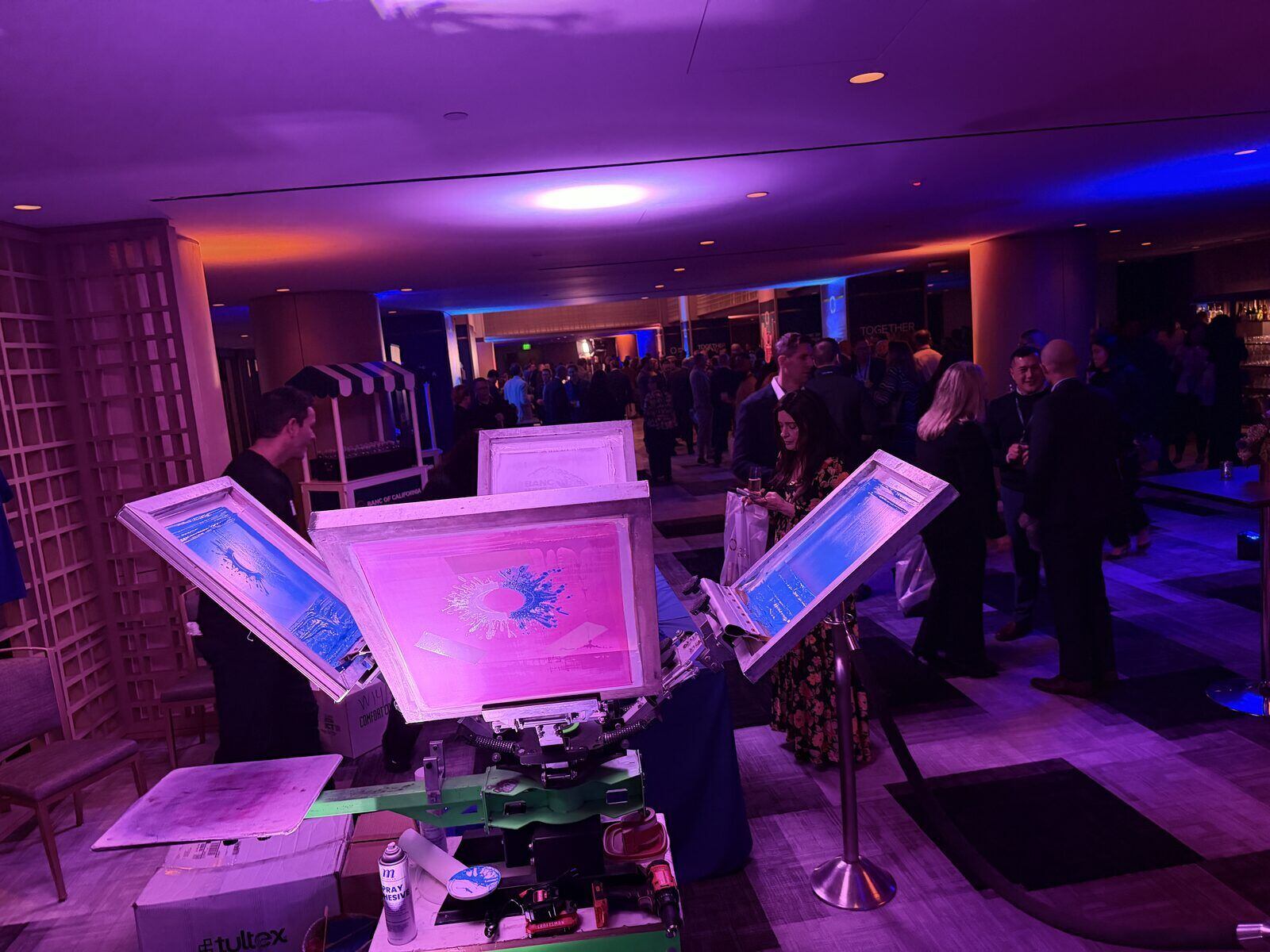 Open screen-print presses set up in a hotel ballroom lit violet and blue, guests mingling in the background