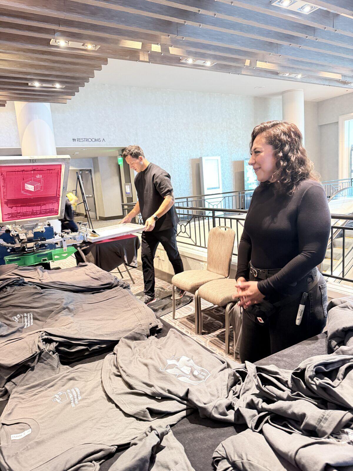 Merch Troop crew setting up a screen-print station and folding finished shirts in a daylit venue